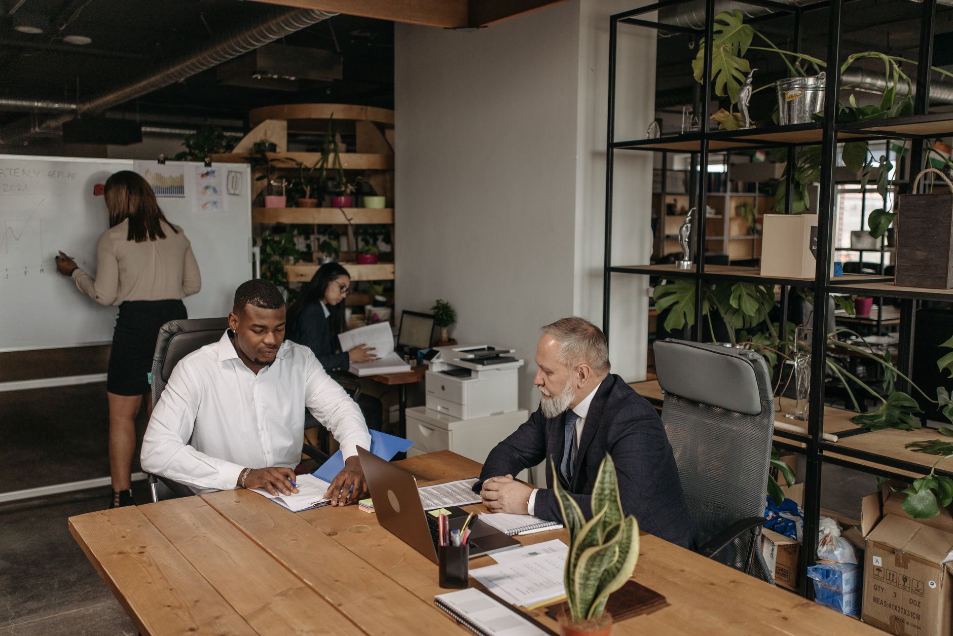 Deux hommes assis à une table en bois avec des documents et un ordinateur portable, pendant qu’une femme écrit sur un tableau blanc dans un bureau.