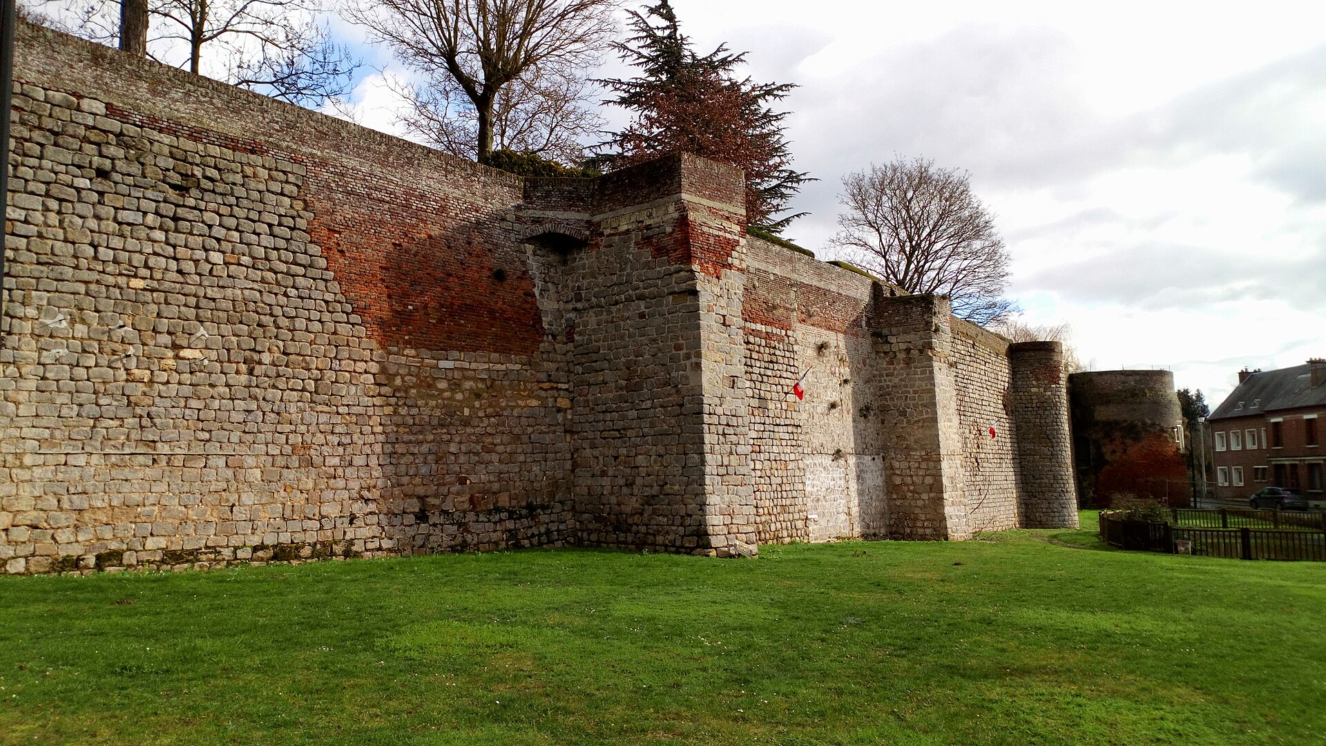 Pan de remparts en pierre et en brique ponctué de tours, au bord d’une pelouse, sous un ciel nuageux.