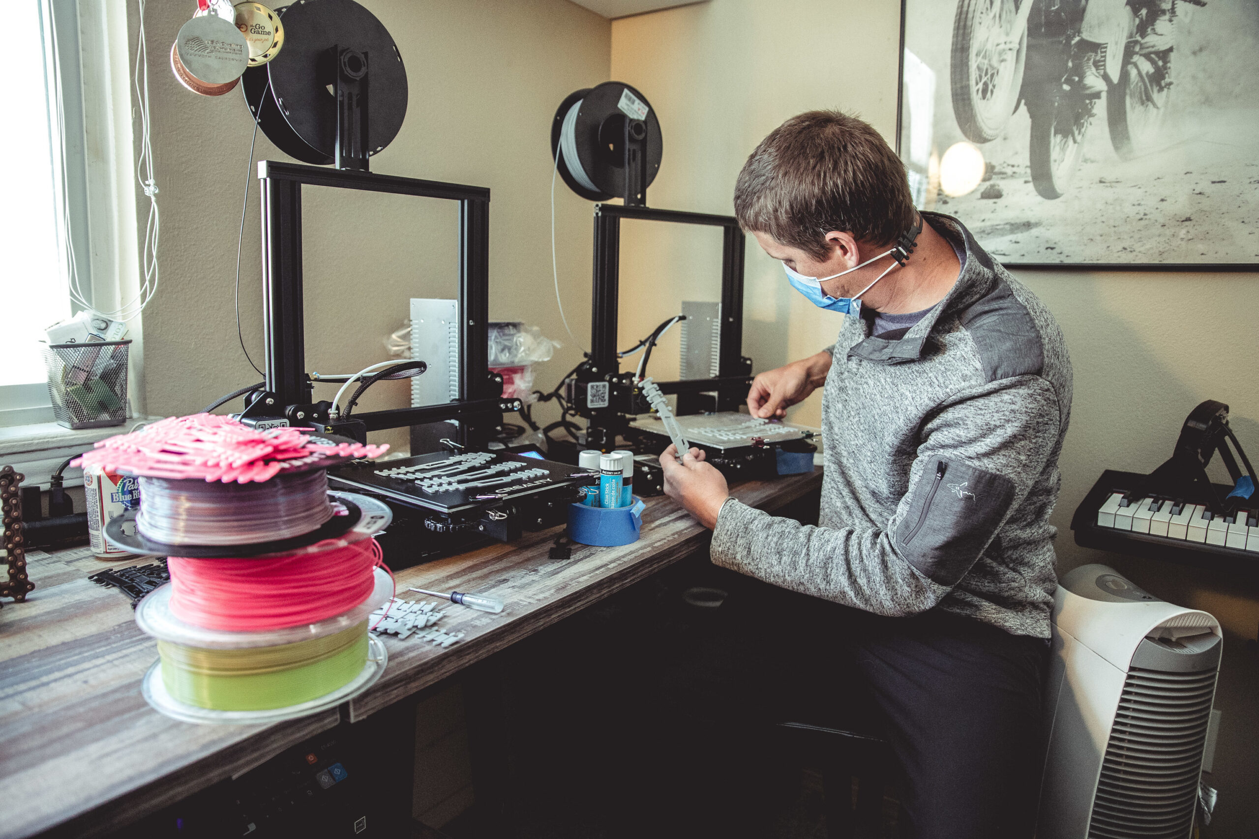 Un homme masqué examine une petite pièce imprimée près de deux imprimantes 3D et de bobines de filament colorées sur un bureau.