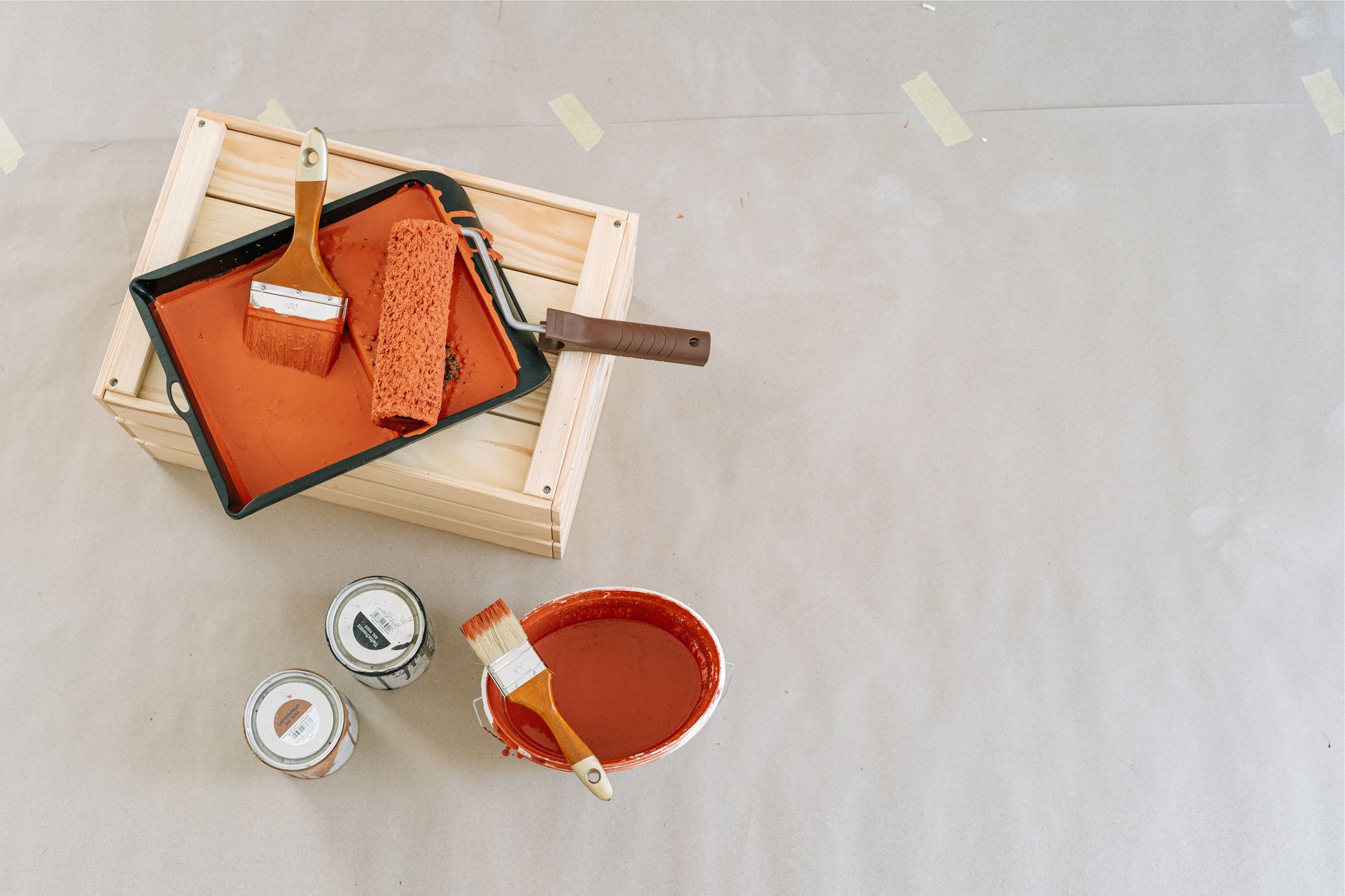 Vue de dessus d’un bac à peinture orange avec rouleau et pinceau sur une caisse en bois, près d’un seau de peinture et de deux pots, sur du papier de protection.